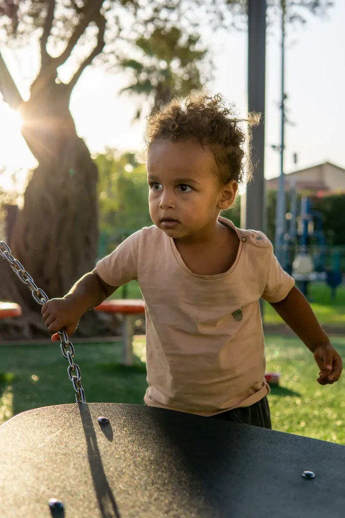 Petit garçon aux cheveux bouclés qui joue avec une balançoire dans un parc en fin de journée.