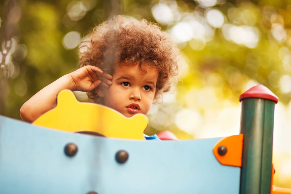 Petit enfant souriant aux cheveux châtains et bouclés se touchant la tête dans une réplique de bateau dans un parc à jeux extérieur